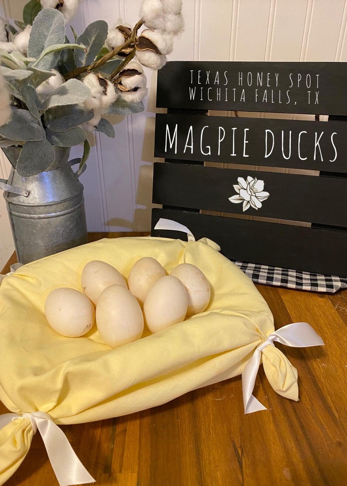 Cream/white magpie duck eggs sitting in a yellow pillow with white bows on a butcher block countertop with cotton in the background, with a black sign that says Magpie Ducks with the Texas Honey Spot logo