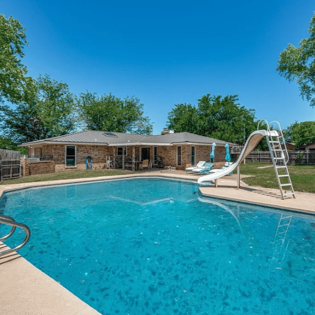 sparkling blue pool with white slide blue skies and green trees in front of a ranch style house